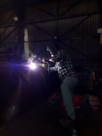 A person wearing a welding helmet is working on a metal structure with a bright welding torch. The surroundings are dark, with industrial equipment and a corrugated metal wall visible in the background.