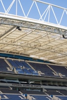 Rows of blue and gray seats in a modern football stadium are positioned under a large, white, lattice-style roof structure. The seating area features the name 'FUTEBOL CLUBE DO PORTO' prominently displayed.