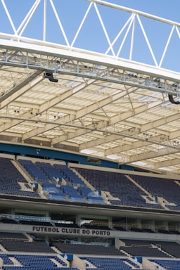 Rows of blue and gray seats in a modern football stadium are positioned under a large, white, lattice-style roof structure. The seating area features the name 'FUTEBOL CLUBE DO PORTO' prominently displayed.