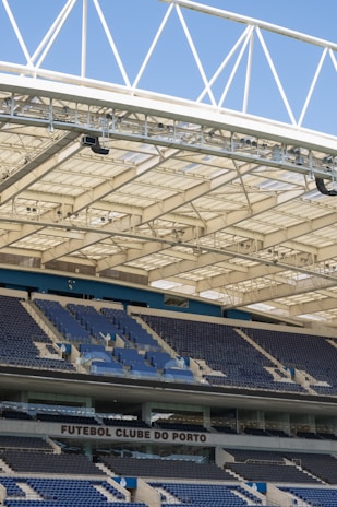 Rows of blue and gray seats in a modern football stadium are positioned under a large, white, lattice-style roof structure. The seating area features the name 'FUTEBOL CLUBE DO PORTO' prominently displayed.