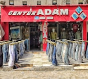 A storefront with a bold red exterior displays the name 'Center Adam Gents Wear' in large, metallic letters. The front features multiple pairs of jeans displayed upside down on mannequins, forming two neat rows leading into the store. Inside, more mannequins showcase various styles of jackets and shirts. A bright and colorful visual setup, emphasizing a casual apparel theme.