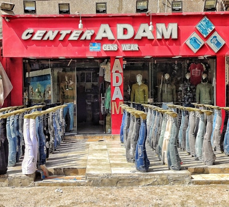 A storefront with a bold red exterior displays the name 'Center Adam Gents Wear' in large, metallic letters. The front features multiple pairs of jeans displayed upside down on mannequins, forming two neat rows leading into the store. Inside, more mannequins showcase various styles of jackets and shirts. A bright and colorful visual setup, emphasizing a casual apparel theme.