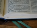 An open book with visible text, placed on a wooden surface. A blue bookmark is seen at the bottom, and a green ribbon is lying alongside it. Lighting highlights the paper texture and the text is well-lit.