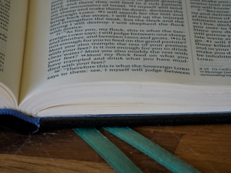 An open book with visible text, placed on a wooden surface. A blue bookmark is seen at the bottom, and a green ribbon is lying alongside it. Lighting highlights the paper texture and the text is well-lit.