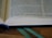 An open book with visible text, placed on a wooden surface. A blue bookmark is seen at the bottom, and a green ribbon is lying alongside it. Lighting highlights the paper texture and the text is well-lit.