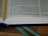 An open book with visible text, placed on a wooden surface. A blue bookmark is seen at the bottom, and a green ribbon is lying alongside it. Lighting highlights the paper texture and the text is well-lit.