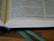 An open book with visible text, placed on a wooden surface. A blue bookmark is seen at the bottom, and a green ribbon is lying alongside it. Lighting highlights the paper texture and the text is well-lit.