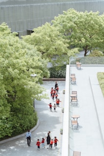 A group of children wearing red and black striped shirts is walking along a pathway surrounded by lush green trees. They are accompanied by a few adults. To the right, there is an open space with square tables and chairs placed along a paved area.