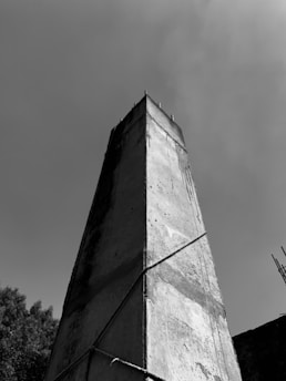 A tall, rectangular concrete structure rises against a clear, cloudless sky. The structure has a rough texture with visible lines and markings, and is reinforced with cables or wires. Part of a tree's foliage can be seen in the lower portion of the image, adding contrast to the starkness of the concrete.