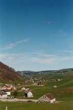 A picturesque rural landscape with rolling green hills dotted with small houses and farm buildings. A lone paraglider is visible high in the clear blue sky, adding a sense of adventure to the serene setting.