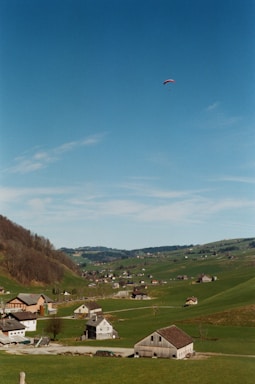 A picturesque rural landscape with rolling green hills dotted with small houses and farm buildings. A lone paraglider is visible high in the clear blue sky, adding a sense of adventure to the serene setting.