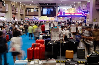 A bustling luggage fair held indoors, with people examining various suitcases displayed on the floor. Bright signage with neon colors advertises the event, while numerous signs and price tags for different brands and models of luggage are visible. The scene is filled with a crowd, creating a busy atmosphere.