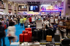 A bustling luggage fair held indoors, with people examining various suitcases displayed on the floor. Bright signage with neon colors advertises the event, while numerous signs and price tags for different brands and models of luggage are visible. The scene is filled with a crowd, creating a busy atmosphere.