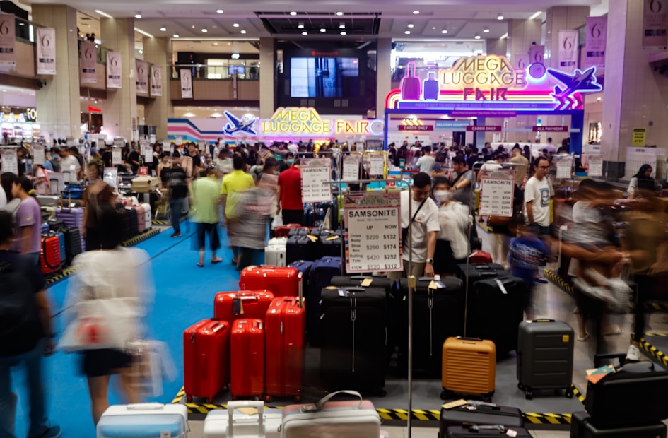 A bustling luggage fair held indoors, with people examining various suitcases displayed on the floor. Bright signage with neon colors advertises the event, while numerous signs and price tags for different brands and models of luggage are visible. The scene is filled with a crowd, creating a busy atmosphere.