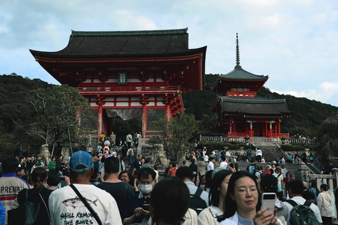 A large group of people gather in front of a traditional Japanese temple with a vibrant red and white gate. The temple features ornate architectural details, surrounded by lush greenery and hills in the background. Visitors are engaged in various activities such as walking and taking photos.