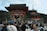 A large group of people gather in front of a traditional Japanese temple with a vibrant red and white gate. The temple features ornate architectural details, surrounded by lush greenery and hills in the background. Visitors are engaged in various activities such as walking and taking photos.