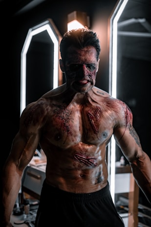 A muscular person with fake severe injuries and makeup giving a gory appearance, standing in front of illuminated mirrors in a dimly lit room. The individual has an aggressive facial expression and is covered in realistic body paint depicting cuts and bruises.