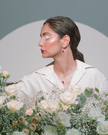 A woman with elegant makeup and jewelry is surrounded by an arrangement of flowers. She is wearing a white outfit and has subtle makeup, with pink lipstick and shimmery eyeshadow. The flowers include white roses and small, delicate blooms against a soft gray background.
