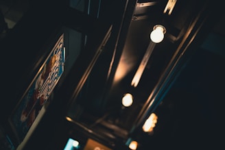 A dimly lit interior displays a series of exposed light bulbs along a dark ceiling. The lights cast a warm glow, creating a cozy atmosphere. A partially visible poster with colorful elements is positioned on the left wall, adding a touch of interest and color contrast.