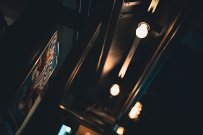A dimly lit interior displays a series of exposed light bulbs along a dark ceiling. The lights cast a warm glow, creating a cozy atmosphere. A partially visible poster with colorful elements is positioned on the left wall, adding a touch of interest and color contrast.