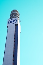 A tall communication tower with a cylindrical structure at the top, featuring a large logo with the letters 'BT' in blue on a white background. The tower is set against a clear blue sky.