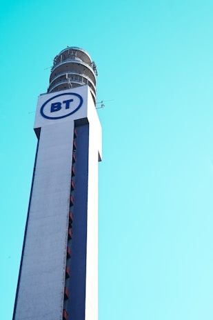 A tall communication tower with a cylindrical structure at the top, featuring a large logo with the letters 'BT' in blue on a white background. The tower is set against a clear blue sky.