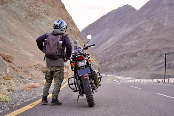A person wearing a helmet and backpack stands beside a motorcycle on a winding mountain road. Rugged mountains and a clear sky form the backdrop, creating a sense of adventure.