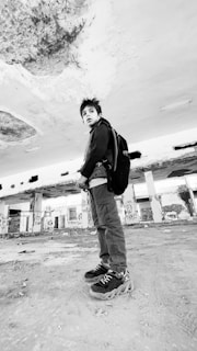 A young person stands in an abandoned building with a worn, exposed ceiling and graffiti-covered walls. The ground is dusty, and the atmosphere seems urban and gritty. The person is wearing a jacket and backpack, looking off into the distance.