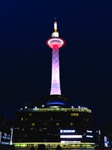 A tall illuminated tower with a striking color gradient from pink to white stands against a dark night sky. The base of the tower emits blue lights, and the surrounding structures are dimly lit with yellow and white lights. The words 'Kyoto Tower Hotel' are visible on the building beneath the tower.