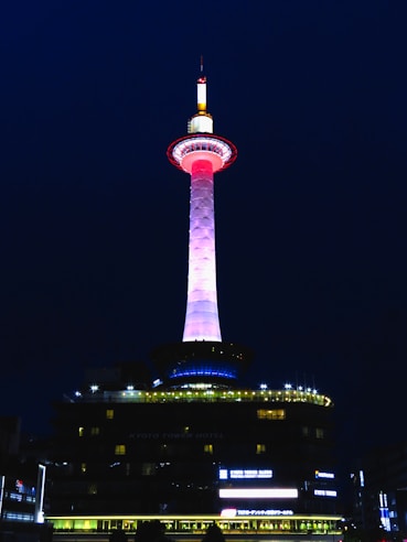 A tall illuminated tower with a striking color gradient from pink to white stands against a dark night sky. The base of the tower emits blue lights, and the surrounding structures are dimly lit with yellow and white lights. The words 'Kyoto Tower Hotel' are visible on the building beneath the tower.