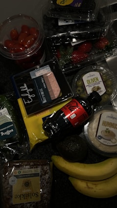 A collection of groceries on a dark surface, including cherry tomatoes in a plastic container, blueberries, strawberries, green olives in a plastic tub, packaged sliced meat, a bottle of Coca-Cola, hummus, a bag of sliced cheese, an avocado, two bananas, a loaf of sunflower seed bread, and a pack of leafy greens.
