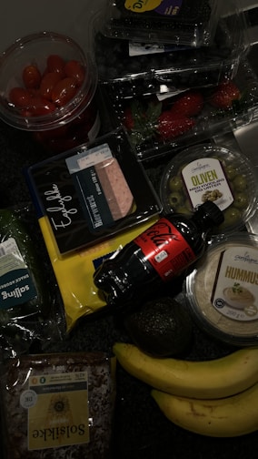 A collection of groceries on a dark surface, including cherry tomatoes in a plastic container, blueberries, strawberries, green olives in a plastic tub, packaged sliced meat, a bottle of Coca-Cola, hummus, a bag of sliced cheese, an avocado, two bananas, a loaf of sunflower seed bread, and a pack of leafy greens.