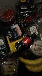 A collection of groceries on a dark surface, including cherry tomatoes in a plastic container, blueberries, strawberries, green olives in a plastic tub, packaged sliced meat, a bottle of Coca-Cola, hummus, a bag of sliced cheese, an avocado, two bananas, a loaf of sunflower seed bread, and a pack of leafy greens.