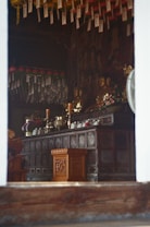 An indoor shrine with intricate wooden carvings and multiple Buddha statues. The shrine is adorned with various offerings, including candles, flowers, and what appears to be incense holders. There are hanging decorations with written prayers.
