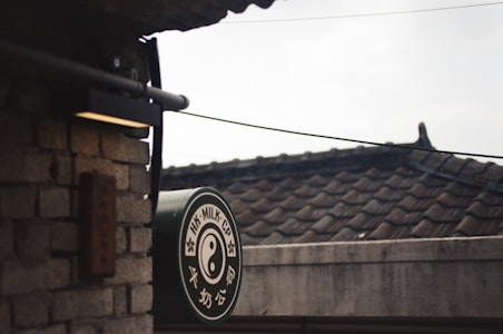 A vintage-style sign with yin-yang symbol and text, mounted on a wall of gray bricks. The background consists of traditional tiled rooftops and overhead cables, creating a nostalgic and slightly rustic atmosphere.