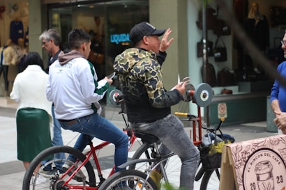 A person wearing a camouflage jacket and cap pedals a bike, operating a wheel grinding tool attached to it. Another person sits beside them on a red bike, looking at a mobile device. Other individuals walk by in the background, and there are storefronts visible. A table with a tablecloth featuring the text 'El Rincon del Cafe' is seen in the foreground.