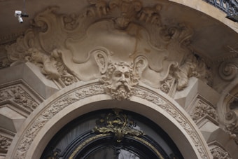 An intricately carved stone facade features an ornate face with flowing hair and a majestic expression. Surrounding the face are elaborate scrolls and leaf designs, adding a sense of grandeur to the architecture. A security camera is mounted on the top left corner.