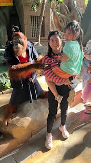 A person is holding a child while standing next to an orangutan dressed in human clothes. The setting appears to be a zoo or an exhibit with a jungle-like backdrop featuring large tree roots and stone structures.