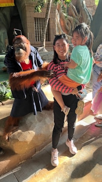 A person is holding a child while standing next to an orangutan dressed in human clothes. The setting appears to be a zoo or an exhibit with a jungle-like backdrop featuring large tree roots and stone structures.