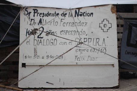 A sign with handwritten text addressed to a president, mentioning waiting for dialogue for two years, and includes dates, a name, and a symbol. Ropes are attached to the sign.