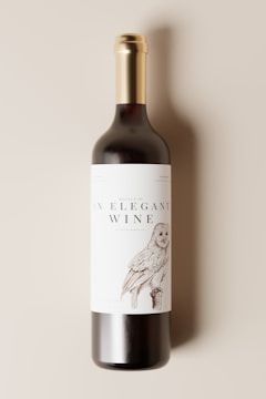 A wine bottle with a gold foil cap and a cream-colored label featuring an illustration of an owl. The label text reads 'An Elegant Wine.' The background is a neutral, soft beige color.