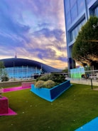 A modern outdoor scene featuring a vibrant and colorful garden area with artificial turf in green, pink, and blue. Tall buildings stand adjacent to the garden, and a stadium with a curved roof is visible in the background. The sky is filled with dramatic clouds lit by soft sunset colors.