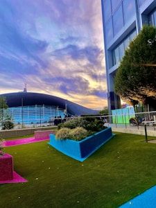 A modern outdoor scene featuring a vibrant and colorful garden area with artificial turf in green, pink, and blue. Tall buildings stand adjacent to the garden, and a stadium with a curved roof is visible in the background. The sky is filled with dramatic clouds lit by soft sunset colors.