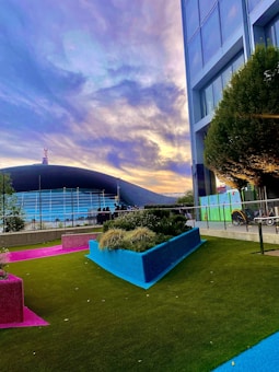 A modern outdoor scene featuring a vibrant and colorful garden area with artificial turf in green, pink, and blue. Tall buildings stand adjacent to the garden, and a stadium with a curved roof is visible in the background. The sky is filled with dramatic clouds lit by soft sunset colors.