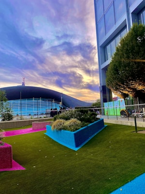 A modern outdoor scene featuring a vibrant and colorful garden area with artificial turf in green, pink, and blue. Tall buildings stand adjacent to the garden, and a stadium with a curved roof is visible in the background. The sky is filled with dramatic clouds lit by soft sunset colors.