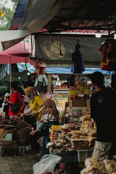 A bustling outdoor market scene featuring several people interacting with various items for sale. The market stalls are filled with packaged goods and fresh produce, and are covered by overhead tarps and umbrellas to provide shade. The environment appears lively with a mix of different colors and textures, contributing to a vibrant and engaging atmosphere.