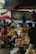 A bustling outdoor market scene featuring several people interacting with various items for sale. The market stalls are filled with packaged goods and fresh produce, and are covered by overhead tarps and umbrellas to provide shade. The environment appears lively with a mix of different colors and textures, contributing to a vibrant and engaging atmosphere.