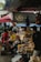 A bustling outdoor market scene featuring several people interacting with various items for sale. The market stalls are filled with packaged goods and fresh produce, and are covered by overhead tarps and umbrellas to provide shade. The environment appears lively with a mix of different colors and textures, contributing to a vibrant and engaging atmosphere.