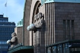 A large stone building with distinctive Art Deco architecture features two prominent sculpted figures holding spherical lamps. The facade is decorated with brick patterns and has vertical lines adding depth to the structure. In the background, modern buildings with glass windows and a flagpole can be seen.