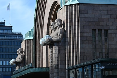 A large stone building with distinctive Art Deco architecture features two prominent sculpted figures holding spherical lamps. The facade is decorated with brick patterns and has vertical lines adding depth to the structure. In the background, modern buildings with glass windows and a flagpole can be seen.
